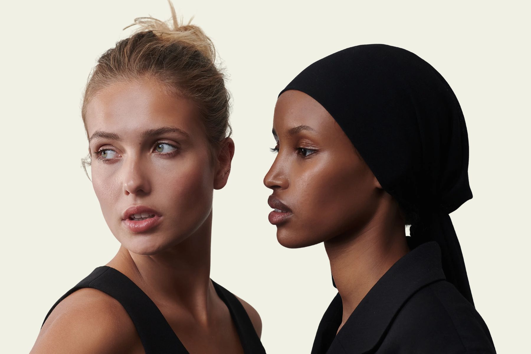 Two women in black clothing pose side by side against a pale background, one with a blonde bun and the other wearing a black hijab, looking in opposite directions.